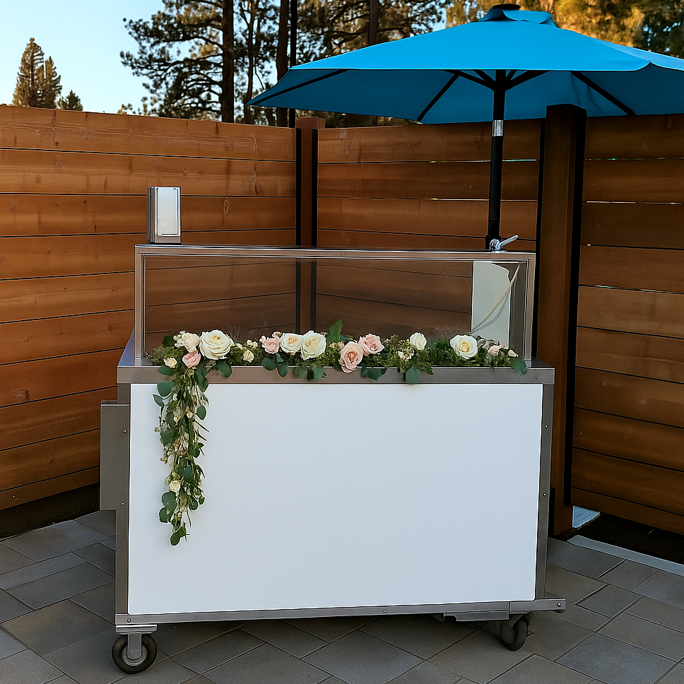 Outdoor setting with a white gelato cart decorated with flowers and a blue umbrella against a wooden fence.