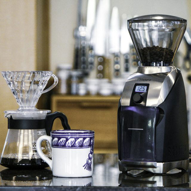 Coffee grinder with a digital display next to a coffee cup and a coffee filter on a countertop.